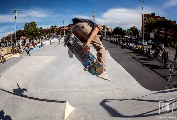 Skater in volo con la tavola in mano durante un trick allo skatepark The Spot di Ostia, pubblico e palazzine sullo sfondo