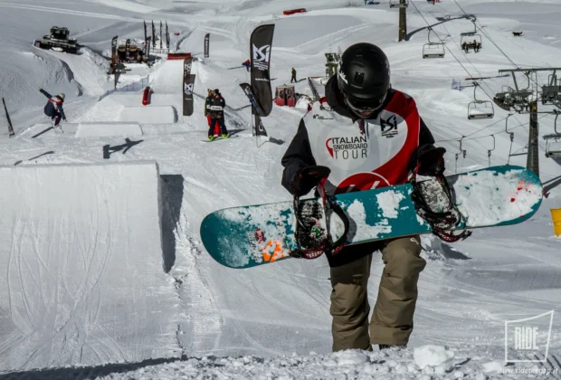Snowboarder con tavola sottobraccio cammina sulla neve durante l'Italian Snowboard Tour, salto e seggiovia sullo sfondo